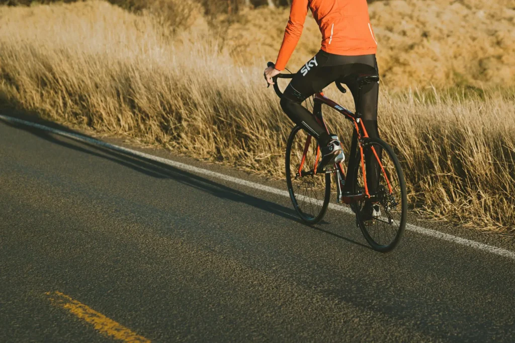 Nell'immagine un ciclista pedala in sella alla sua bicicletta arancio e nera su una strada asfaltata con accanto un prato. L'uomo indossa abbagliamento tecnico con pantaloni neri e maglietta arancione.