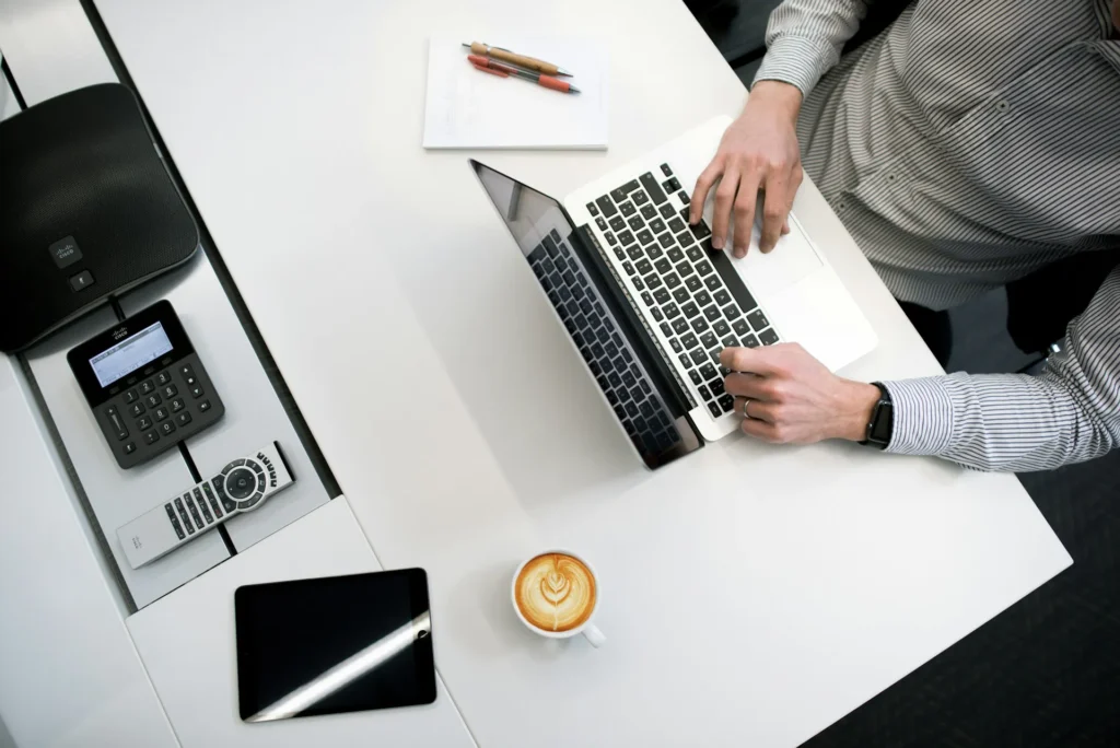 Nell'immagine un uomo in camicia a righe lavora a un laptop posato su una scrivania bianca. Sulla scrivania ci sono anche: accanto al pc un notebook e 2 penne, un cappuccino e un tablet; sulla sinistra un telecomando, uno speaker e un monitor con tastiera.